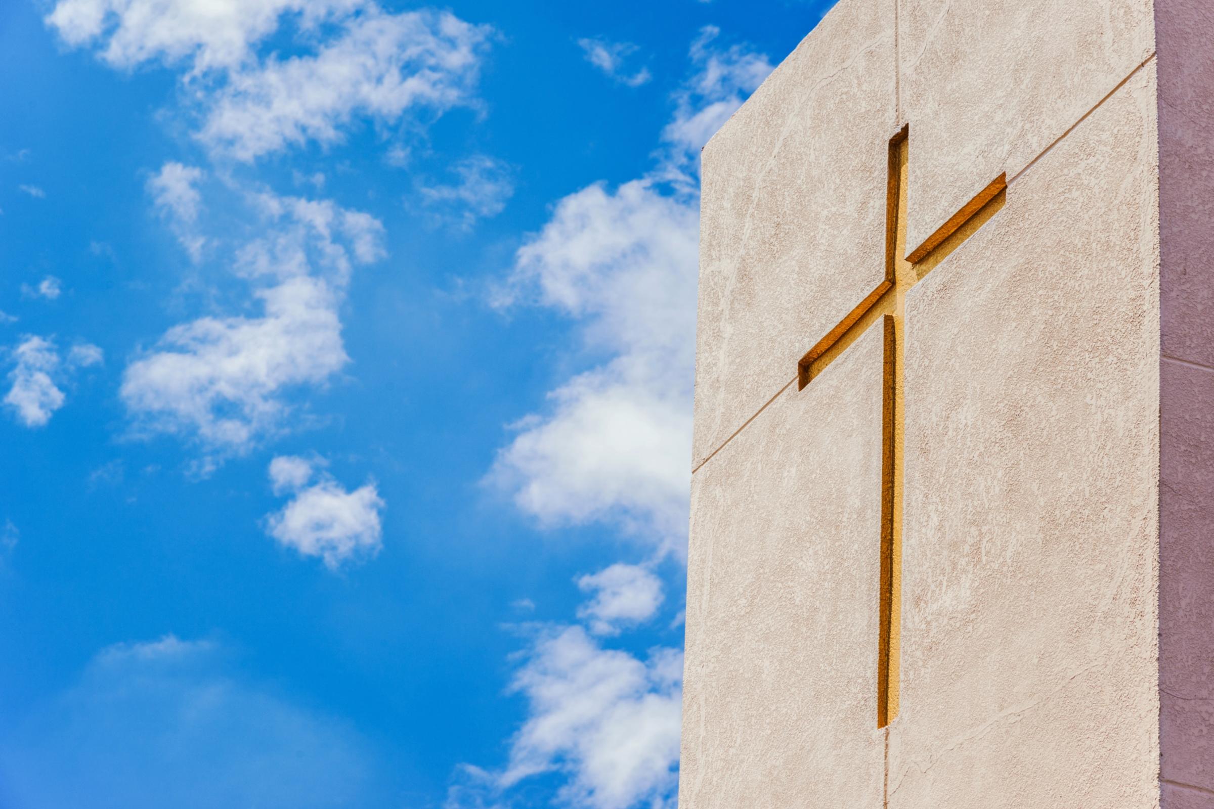 Betonfassade mit eingelassenem goldenem Kreuz, aufgenommen gegen einen leicht bewölkten strahlend blauen Himmel