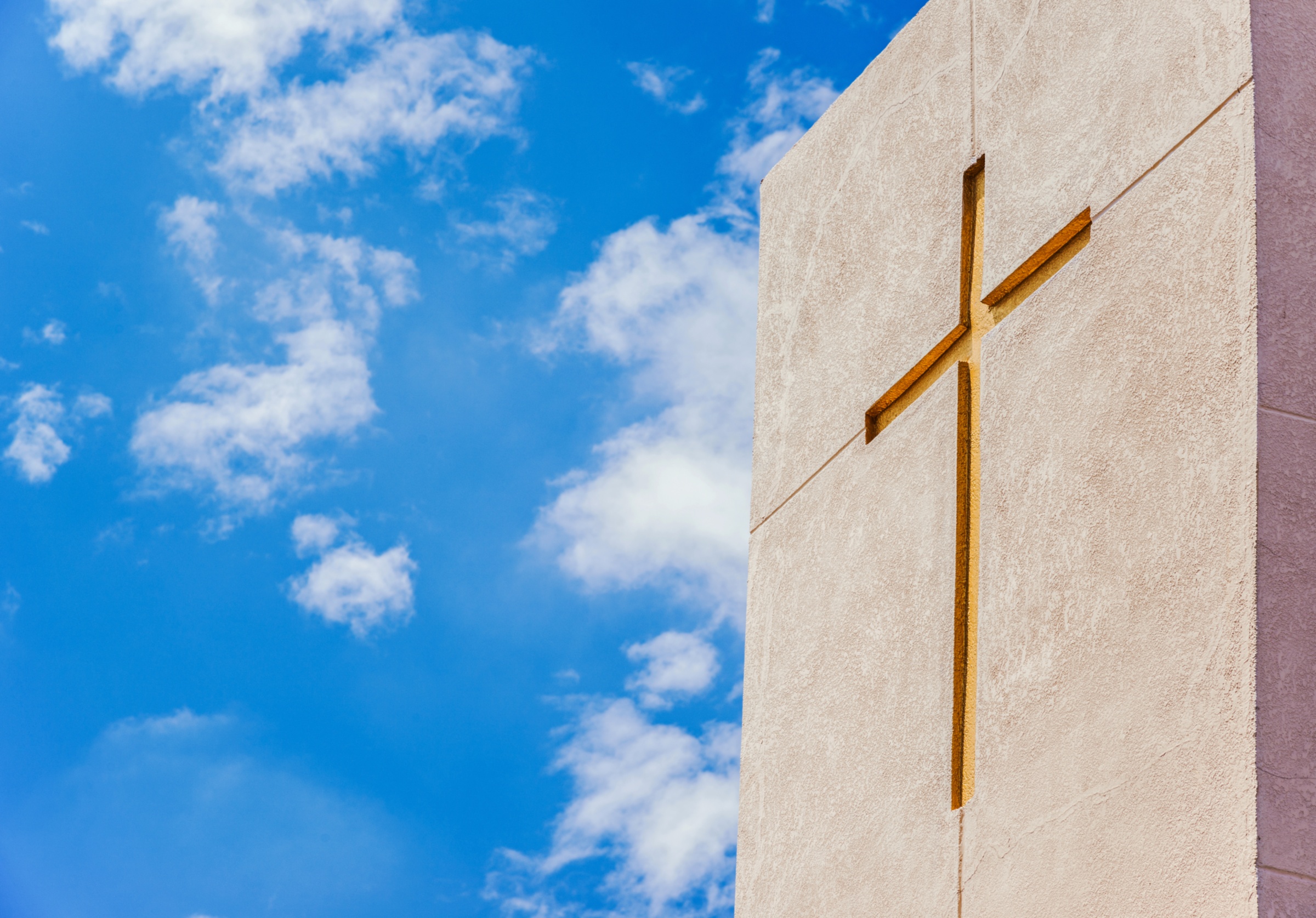 Betonfassade mit eingelassenem goldenem Kreuz, aufgenommen gegen einen leicht bewölkten strahlend blauen Himmel