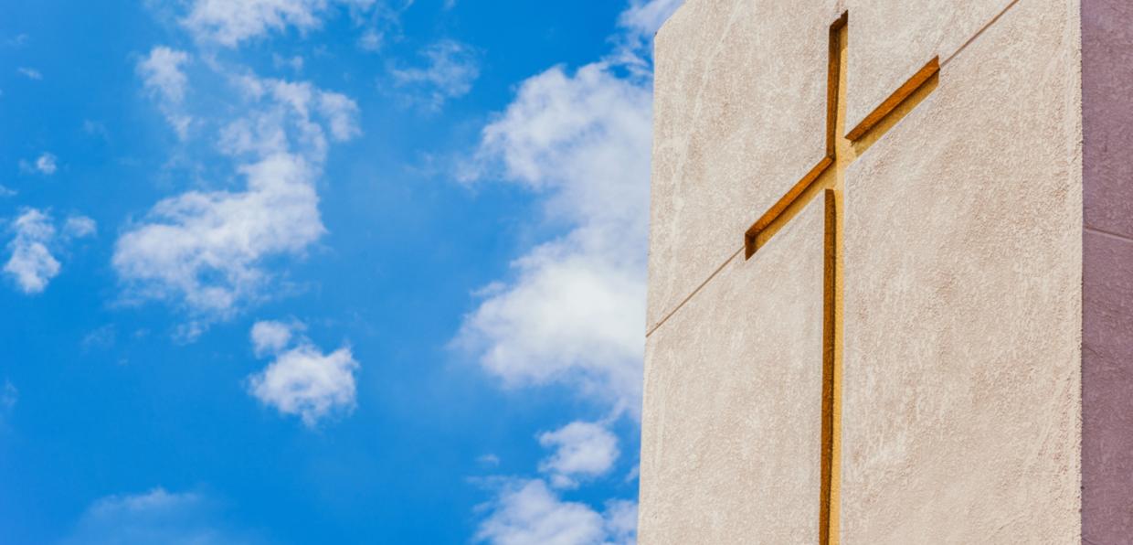 Betonfassade mit eingelassenem goldenem Kreuz, aufgenommen gegen einen leicht bewölkten strahlend blauen Himmel