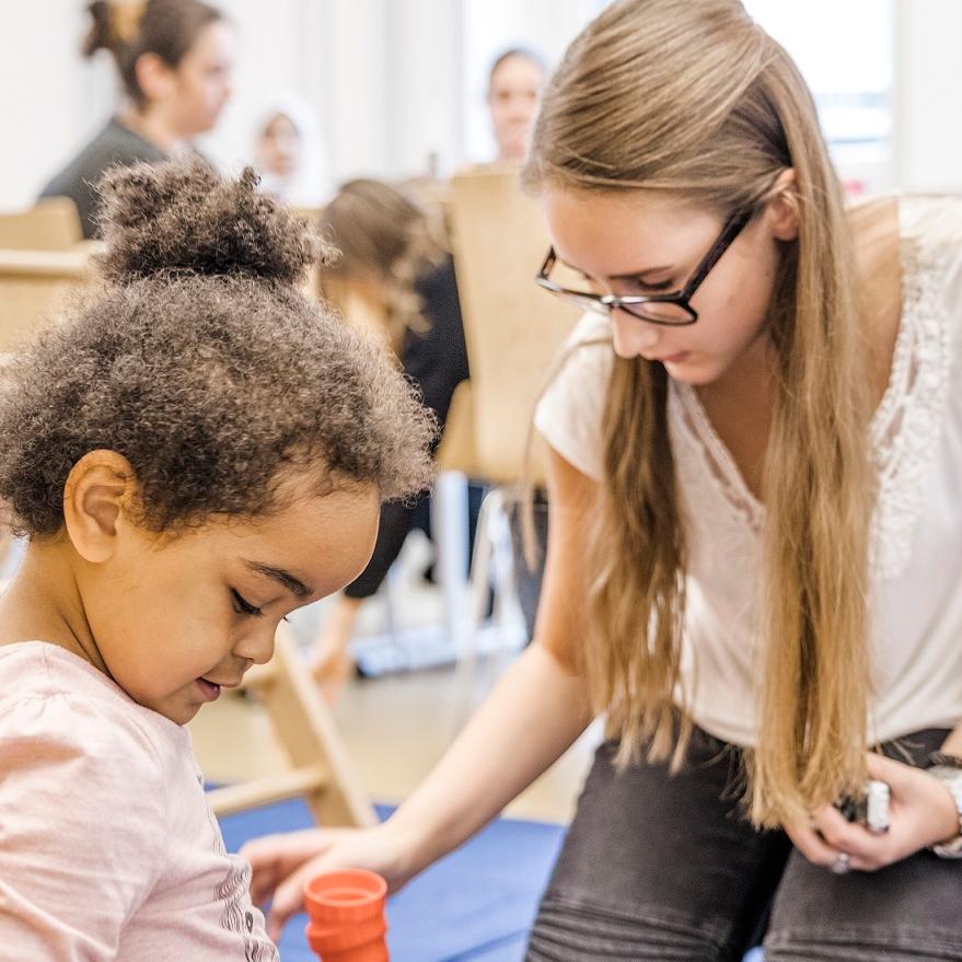 Eltern und KInder im Eltern-Kind-Kurs, im Vordergrund eine Mutter mit Kindern beim Spiel
