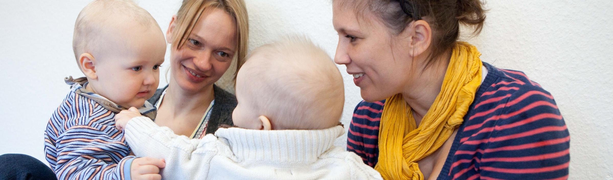Zwei Mütter mit ihren Babies auf dem Schoß schauen interessiert zu, wie ihre Babies im Eltern-Kinder-Kurs aufeinander reagieren.