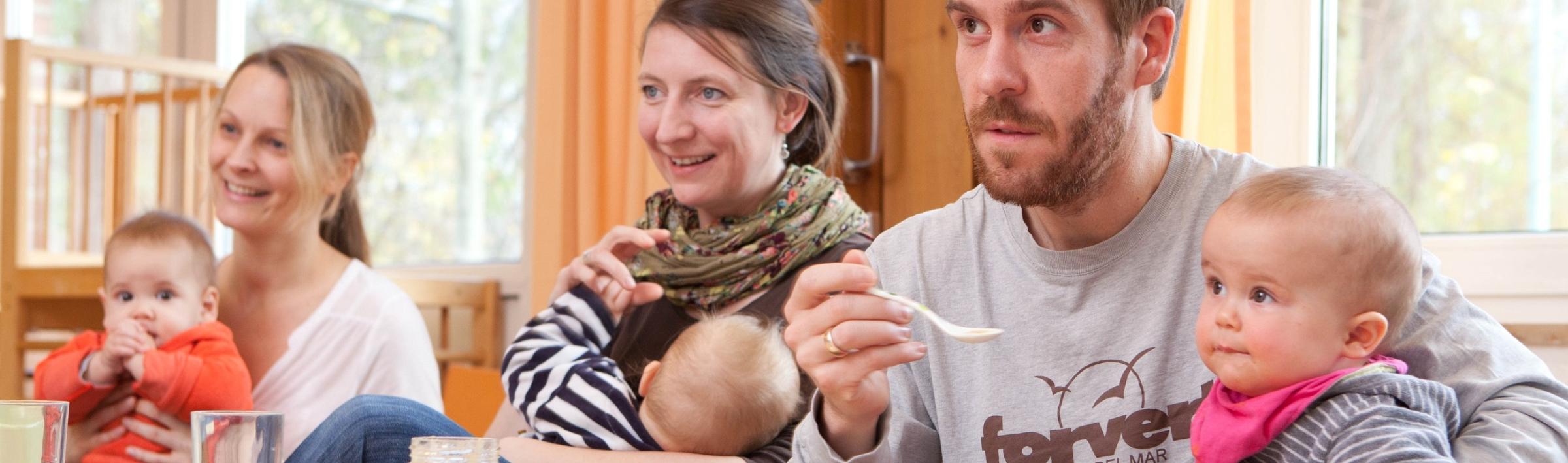 Väter und Mütter mit ihren Kindern auf dem Schoß hören im Eltern-Kind-Kurs aufmerksam zu.
