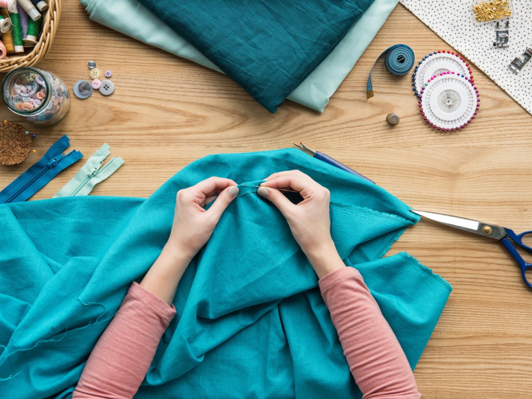 shutterstock_765818908_LightField Studios_Hände_nähen_Stoffe_blau_Faden_Nähzubehör_Schere_Nadel_Reißverschluss_Nadelkissen_Knöpfe_Maßband_frei