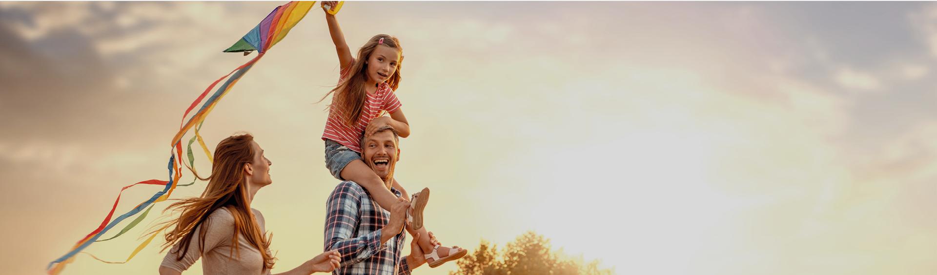 Eine Familie läuft fröhlich über eine weite Wiese und lässt einen Drachen steigen.