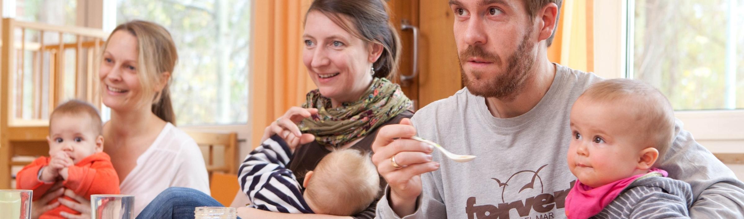 Väter und Mütter mit ihren Kindern auf dem Schoß hören im Eltern-Kind-Kurs aufmerksam zu.